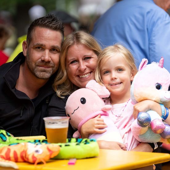 Eine Familie aus drei Personen, ein Mann, eine Frau und ein junges Mädchen, lächelt und posiert für ein Foto mit Kuscheltieren auf einem Tisch.