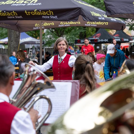 Eine Frau in einer roten Weste dirigiert eine Brassband bei einer Veranstaltung im Freien unter einem Regenschirm.