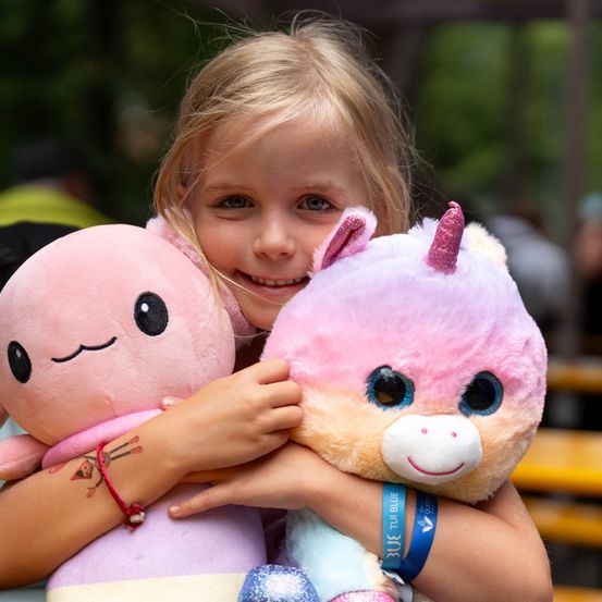 Ein junges Mädchen mit blonden Haaren hält zwei rosa Plüschtiere, eines mit einem Einhornhorn und das andere mit einem lächelnden Gesicht. Sie trägt ein blaues Band mit Text an ihrem Arm.