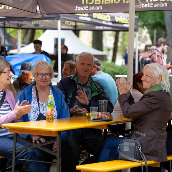 Eine Gruppe von Menschen, darunter ältere Erwachsene, sitzt unter einem Zelt, klatscht und lächelt. Vor ihnen steht ein Tisch mit einer Flasche und Gläsern.