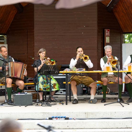 Eine Gruppe von Musikern in traditioneller Kleidung spielt auf einer Bühne mit verschiedenen Instrumenten. Eine Frau spielt Akkordeon, während drei Männer Trompeten spielen. Sie sitzen auf einer Bank mit einem Tisch davor.