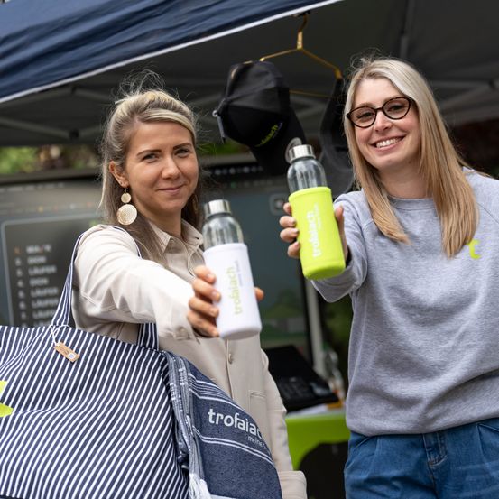 Zwei Frauen lächeln und halten grüne Flaschen unter einem Zelt. Eine Frau hält eine weiße Flasche, die andere eine grüne. Sie stehen neben einer gestreiften Tasche und einem schwarzen Hut.