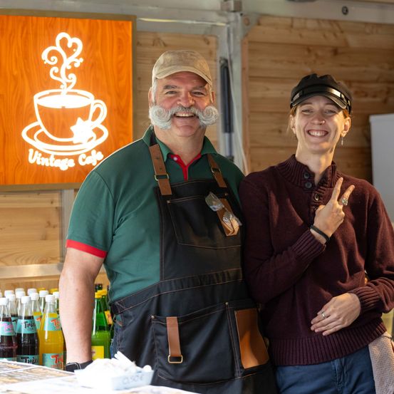 Ein Mann und eine Frau stehen vor einem Vintage-Cafe-Schild, lächeln und posieren für ein Foto. Der Mann trägt ein grünes Shirt und eine Schürze, die Frau ein kastanienbraunes Sweatshirt und eine Mütze. Dahinter ist eine Holzwand und Flaschen mit Limonade zu sehen.