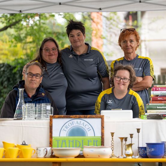 Fünf Personen in passenden grauen und gelben Shirts stehen hinter einem Tisch mit verschiedenen Artikeln zum Verkauf. Der Tisch ist mit einem weißen Tuch bedeckt und hat ein Schild, das 'Trofaiach Tandier' steht. Es gibt Tassen, Teller und andere Gegenstände auf dem Tisch. Die Leute lächeln und posieren für ein Foto.
