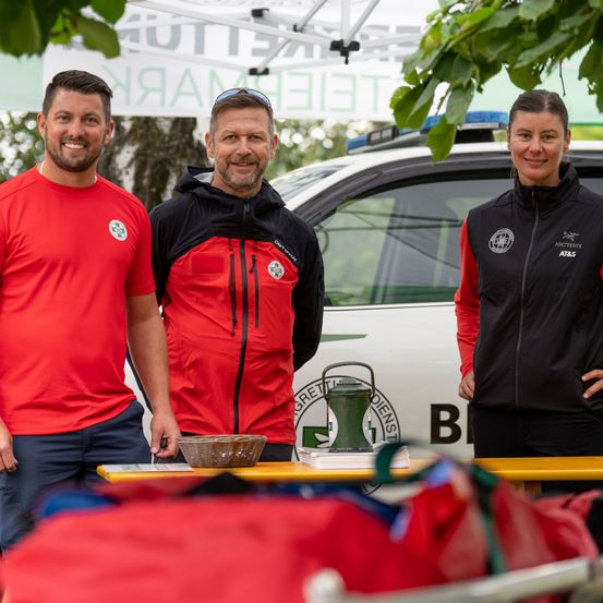 Drei Personen, zwei Männer und eine Frau, stehen vor einem weißen Fahrzeug und lächeln für ein Foto. Sie tragen passende rote und schwarze Jacken mit Logos. Ein Tisch mit einem Korb und einer Laterne steht vor ihnen.