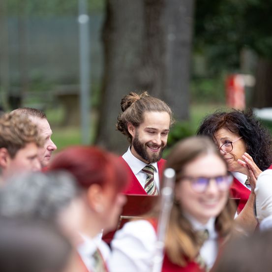 Eine Gruppe von Menschen, wahrscheinlich Musiker, versammelt sich in einem Park. Ein Mann in der Mitte lächelt, während andere zuschauen.
