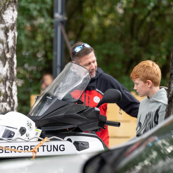Ein Mann in einer rot-schwarzen Jacke steht neben einem Motorrad mit einem Helm darauf. Ein junger Junge mit roten Haaren schaut das Motorrad an.