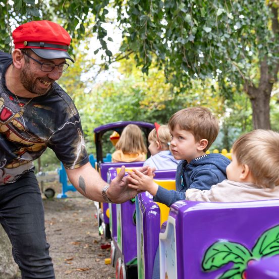 Ein Mann mit rotem Hut und Brille hält die Hand eines kleinen Jungen auf einer bunten Zugfahrt im Park.
