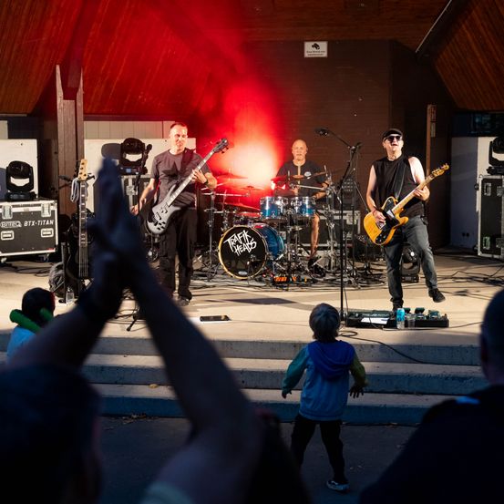 Eine Rockband spielt auf der Bühne mit roter Beleuchtung und begeistert ein junges Publikumsmitglied im Vordergrund.