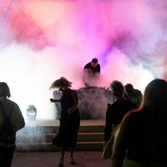 Ein DJ spielt Musik auf der Bühne, während eine Frau vor ihm tanzt, umgeben von Rauch und einer Menge Leute.