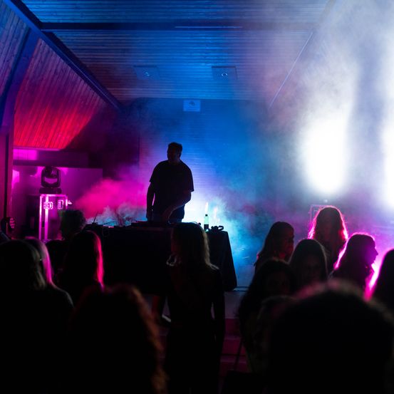 Ein DJ steht hinter einem Tisch in einem Club, umgeben von einer Menge unter leuchtenden blauen und rosafarbenen Lichtern.