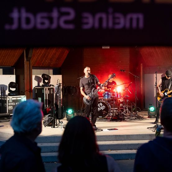 Eine Band spielt auf der Bühne mit roter Beleuchtung und einem Banner mit der Aufschrift '2552 niam'. Der Leadsänger spielt den Bass, während ein anderer Bandmitglied das Schlagzeug spielt.