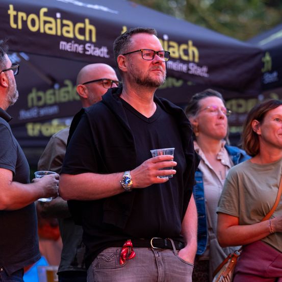 Eine Gruppe von Menschen steht unter einem Zelt, ein Mann in der Mitte hält ein Getränk. Der Mann trägt eine Brille und eine Uhr.