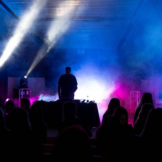 Ein DJ steht auf der Bühne mit einem Tisch vor ihm, umgeben von farbenfrohen Bühnenlichtern und einer Menge von Menschen.