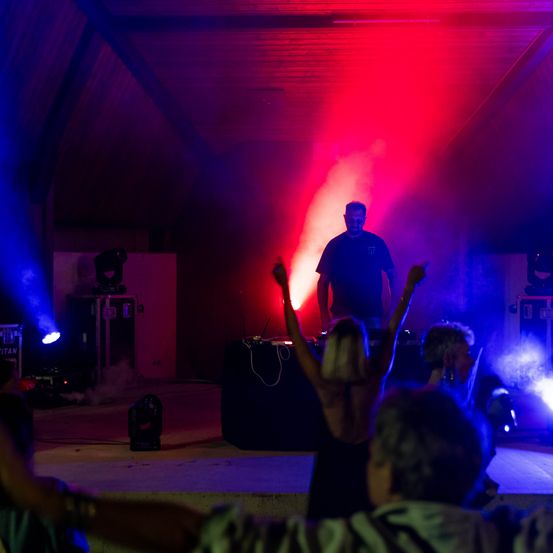 Ein Mann steht auf der Bühne mit einer DJ-Anlage, umgeben von einer Menge tanzender Menschen unter roten und blauen Bühnenlichtern.