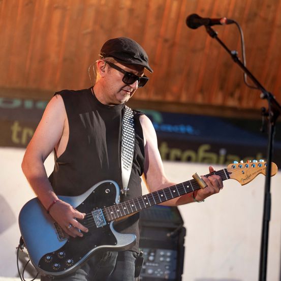 Ein Mann in einem schwarzen ärmellosen Shirt und Hut spielt eine blaue E-Gitarre auf der Bühne.