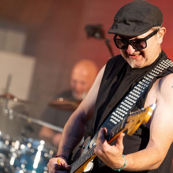 Ein Mann in einem schwarzen Hut und Sonnenbrille spielt Gitarre auf der Bühne, ein Schlagzeuger steht dahinter.