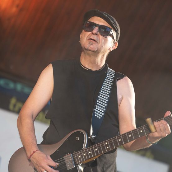 Ein Mann in einem schwarzen ärmellosen Shirt und einem Hut spielt eine E-Gitarre auf der Bühne.