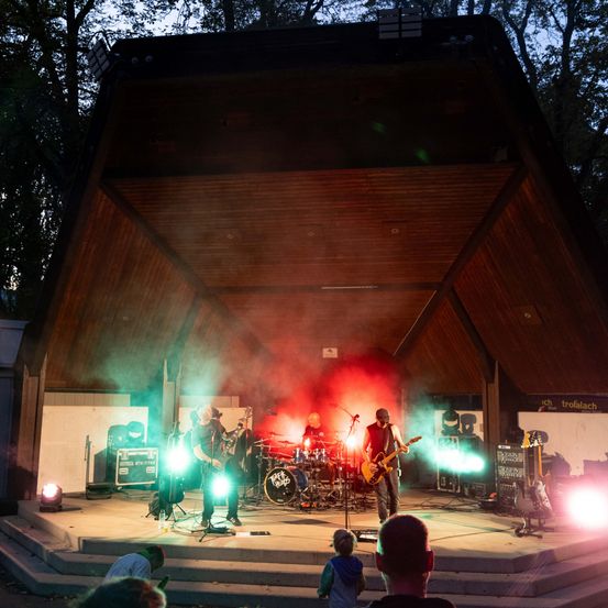 Eine Rockband spielt auf einer Bühne mit roten und grünen Lichtern unter einem hölzernen Dach, während ein Publikum zuschaut.