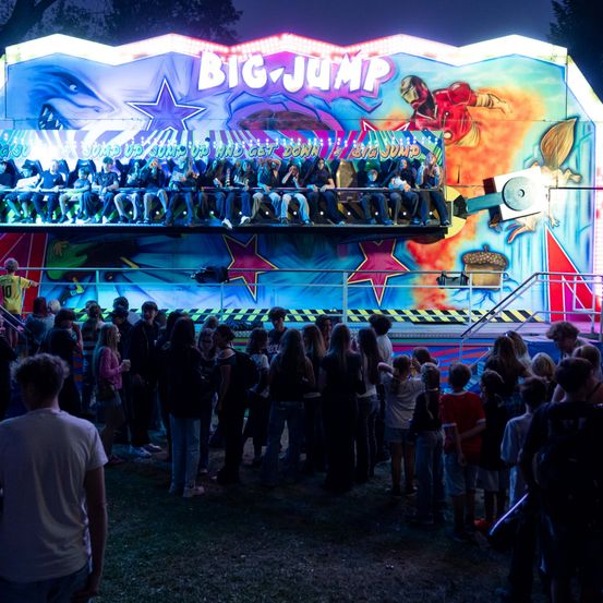 Eine Menschenmenge steht vor einem hell beleuchteten 'Big Jump'-Karussell, mit sitzenden Fahrgästen und der farbenfrohen, dynamischen Kunst des Karussells sichtbar.