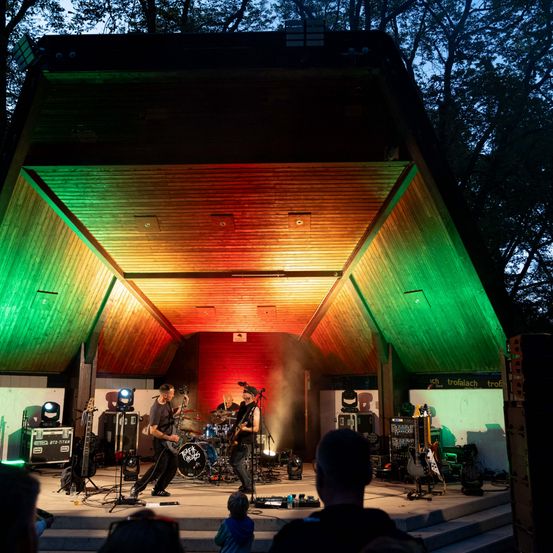 Eine Band spielt auf einer Bühne mit einer leuchtenden, mehrfarbigen Decke, unter einem klaren Abendhimmel. Das Publikum sitzt vor der Bühne und beobachtet die Aufführung.