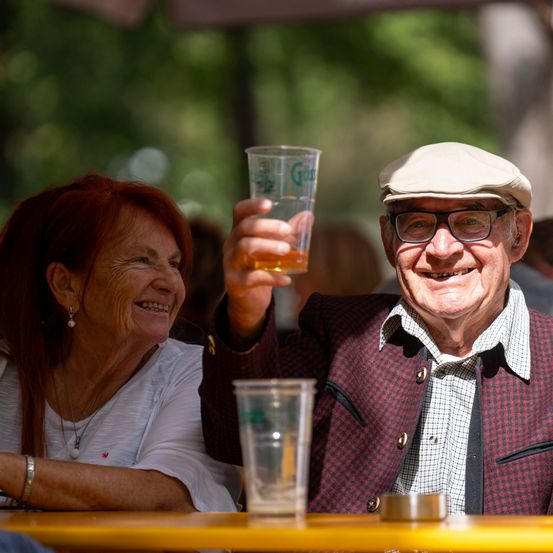 Ein älteres Paar sitzt an einem Tisch, der Mann hält ein Glas Bier in der Hand und lächelt, während die Frau neben ihm zusieht.