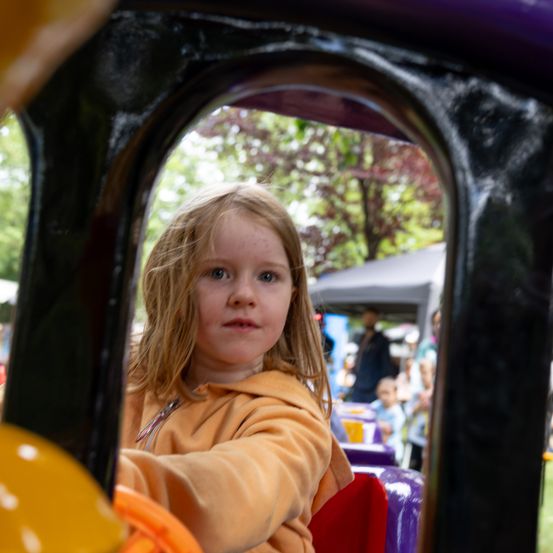 Ein kleines Mädchen mit blonden Haaren sitzt in einem farbenfrohen, spielzeugartigen Fahrzeug, wahrscheinlich auf einem Jahrmarkt, mit einem verschwommenen Hintergrund von Menschen und Bäumen.