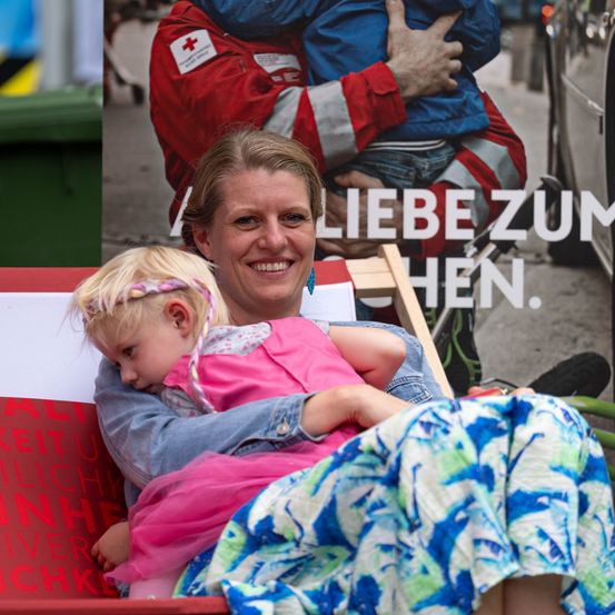 Eine lächelnde Frau hält ein schlafendes Kind auf dem Schoß vor einem Plakat mit einem Rotkreuz-Helfer.