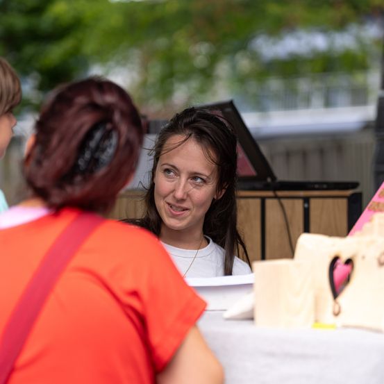 Eine Frau mit dunklem Haar lächelt eine andere Frau in einem orangefarbenen Shirt an, mit einem Holztisch und einem herzförmigen Objekt im Hintergrund.