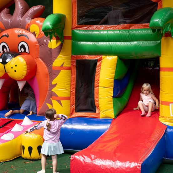 Zwei kleine Mädchen spielen in einem bunten aufblasbaren Löwen-Bounce-Haus. Ein Mädchen rutscht die Rutsche hinunter, während das andere zuschaut.
