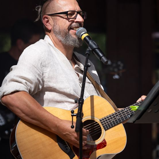 Ein Mann mit Bart und Brille spielt Gitarre und singt in ein Mikrofon. Er trägt ein weißes Hemd und hat eine Gitarrenschlaufe über der Schulter.