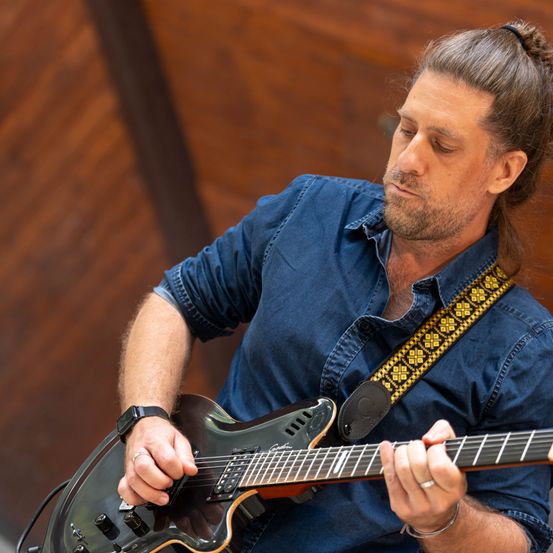 Ein Mann mit Bart und Pferdeschwanz spielt Gitarre und schaut nach unten. Er trägt ein blaues Hemd und eine schwarze Armbanduhr.