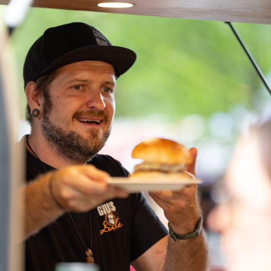 Ein Mann mit Bart und schwarzer Kappe hält ein Sandwich in einem Food Truck.