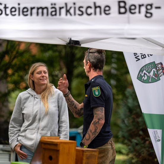 Ein tätowierter Mann spricht mit einer Frau unter einem Zelt mit einem Schild, das 'Steiermarkische Berg' besagt.