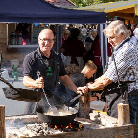 Zwei Männer kochen Essen über einem offenen Feuer bei einer Veranstaltung im Freien. Ein Mann wirft das Essen in einer Pfanne, während der andere einen Kochlöffel hält. Sie sind von einer Menge umgeben.