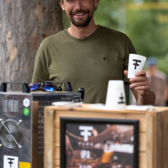 Ein Mann in einem grünen T-Shirt hält einen Becher mit dem Buchstaben F und lächelt vor einem Holztresen.