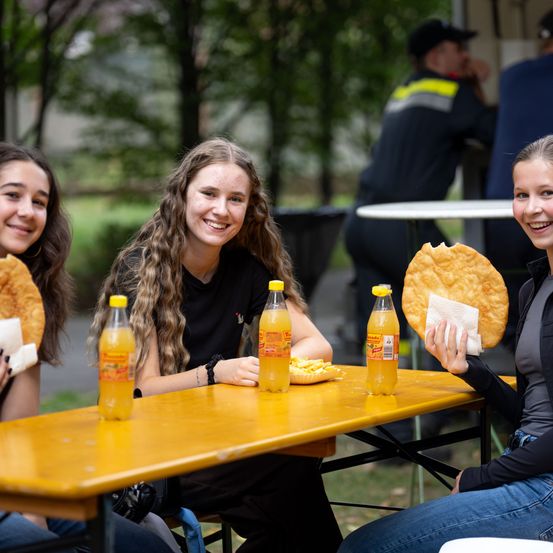 Drei junge Frauen sitzen an einem Tisch im Freien, lächeln und halten Essen. Flaschen mit Orangensoda stehen auf dem Tisch. Ein Mann sitzt im Hintergrund an einem anderen Tisch.