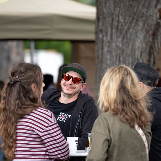 Ein Mann mit Sonnenbrille und grüner Kappe sitzt an einem Tisch mit drei Frauen, von denen eine lange blonde Haare hat. Der Mann trägt ein schwarzes Shirt mit weißem Text.
