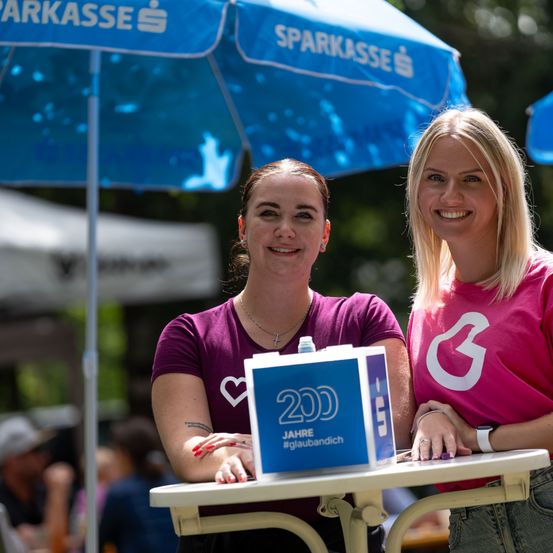 Zwei Frauen stehen unter einem blauen Schirm und lächeln für ein Foto. Eine hält eine Box mit der Aufschrift '200 Jahre #glaubandich'.