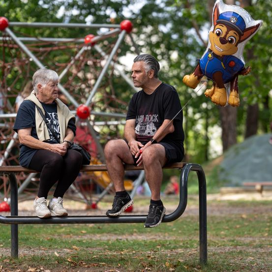 Eine ältere Frau und ein Mann sitzen auf einer Parkbank, mit einem Luftballon eines Cartoon-Hundes, der über ihnen schwebt.