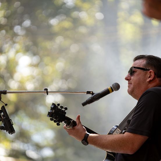 Ein Mann spielt Gitarre und singt in ein Mikrofon auf der Bühne. Er trägt eine Sonnenbrille und ein schwarzes T-Shirt.