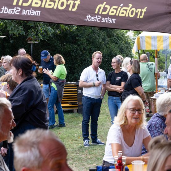 Eine Gruppe von Menschen versammelt sich im Freien, wahrscheinlich bei einem Festival. Einige stehen, andere sitzen und alle sind in Gespräche vertieft. Ein Mann in einem weißen Hemd hält ein Glas Bier, während eine Frau in einem grünen Shirt ein Glas Wein hält.