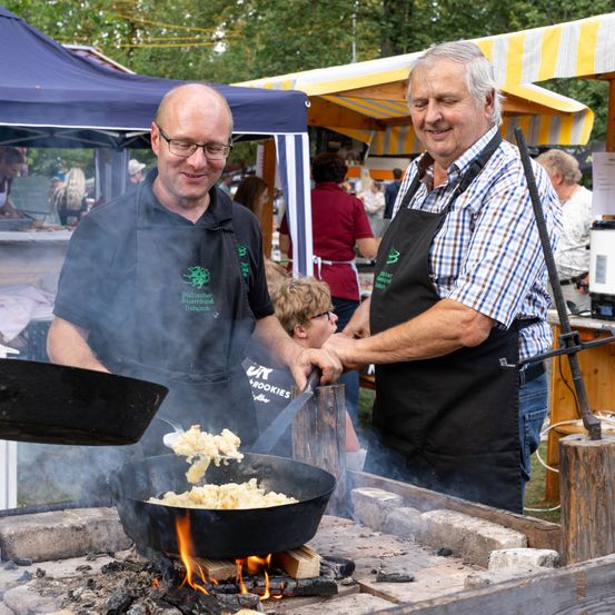 Zwei Männer in Schürzen kochen Essen über einem offenen Feuer auf einem Outdoor-Markt. Ein Mann hält eine Schöpfkelle, während der andere hilft.