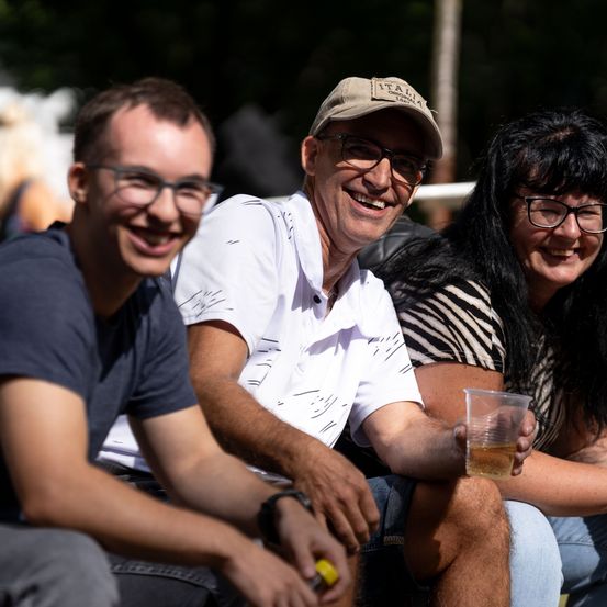 Drei Menschen sitzen zusammen und lächeln. Ein Mann in einem weißen Hemd hält ein Getränk, während eine Frau in einem Zebra-Print-Shirt neben ihm sitzt. Ein weiterer Mann mit Brille sitzt links.