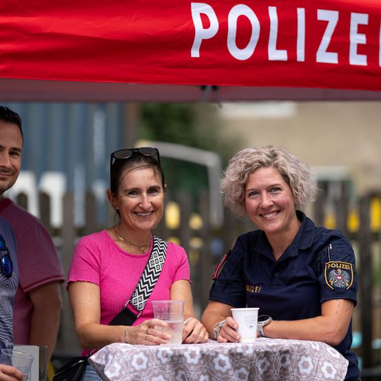Zwei Frauen sitzen an einem Tisch unter einem roten Zelt mit der Aufschrift 'POLIZEI'. Eine trägt ein rosa Shirt und die andere eine blaue Uniform. Beide halten Tassen und lächeln. Ein Mann steht hinter ihnen.
