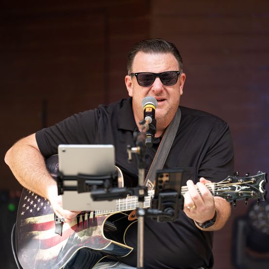 Ein Mann in einem schwarzen Shirt und Sonnenbrille spielt eine akustische Gitarre mit amerikanischem Flaggendesign und singt in ein Mikrofon auf der Bühne.