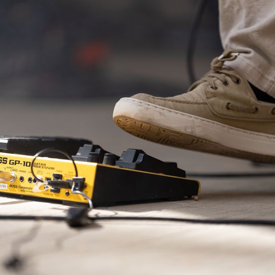 Ein Fuß steht auf einem gelben und schwarzen Boss GP-10 Gitarrenprozessor-Pedal mit angeschlossenen Kabeln.