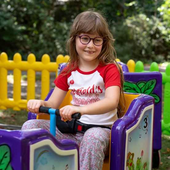 Ein junges Mädchen mit Brille fährt in einem Park mit einem bunten Spielzeugzug. Sie lächelt und hält das Lenkrad. Der Zug hat die Nummer 3 auf der Seite. Dahinter steht ein gelber Zaun.