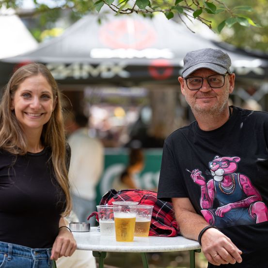 Ein Mann und eine Frau sitzen an einem Tisch mit drei Biergläsern und lächeln für ein Foto. Der Mann trägt ein schwarzes T-Shirt mit einem rosafarbenen Katzenmotiv, und die Frau trägt ein schwarzes Shirt und Jeans. Im Hintergrund ist eine verschwommene Umgebung mit einem Zelt und Menschen.
