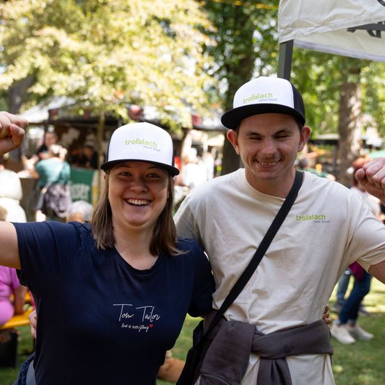 Zwei Menschen lächeln und posieren für ein Foto bei einer Outdoor-Veranstaltung. Beide tragen weiße und schwarze Mützen mit dem Wort 'trofaiaach' darauf. Die Frau trägt ein blaues T-Shirt mit dem Text 'Tom Tailor' und einem Herzsymbol. Der Mann trägt ein weißes T-Shirt mit einem schwarzen Riemen. Hinter ihnen sind verschwommene Menschen und Bäume.